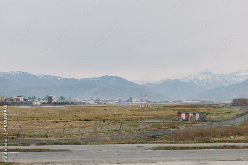 Naklejka premium Empty rural landscape with overcast sky and distant mountains, featuring grassy fields and small buildings near a paved road. Concept of countryside, nature, and calm environment.