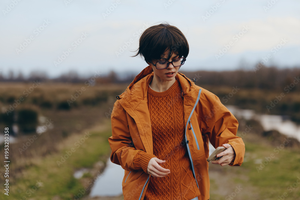 Fototapeta premium Young woman lifestyle outdoors wearing orange jacket and sweater walks on rural path with puddles. Autumn season, casual fashion and nature concept with overcast sky background.