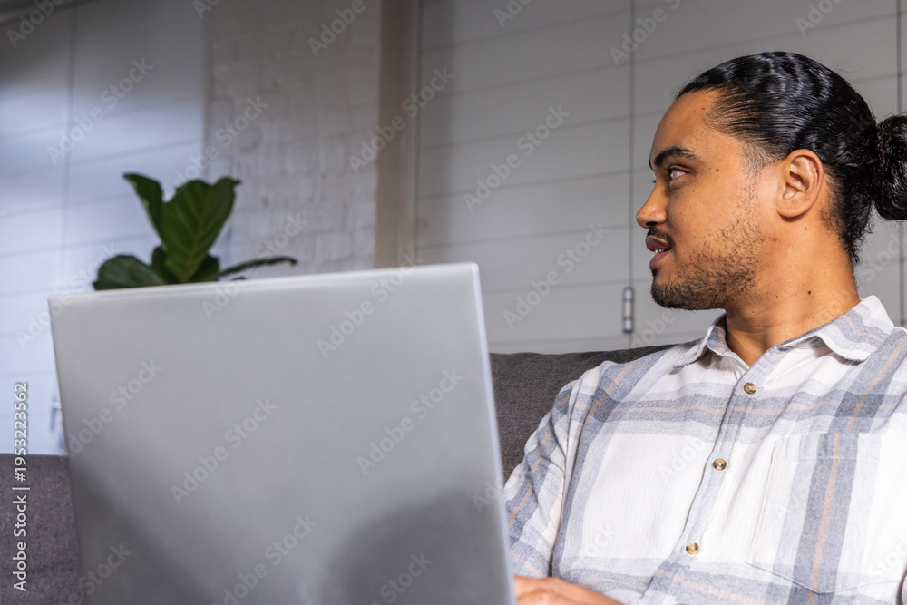 Fototapeta premium Asian man sitting on gray sofa in living room, typing on silver laptop, copy space