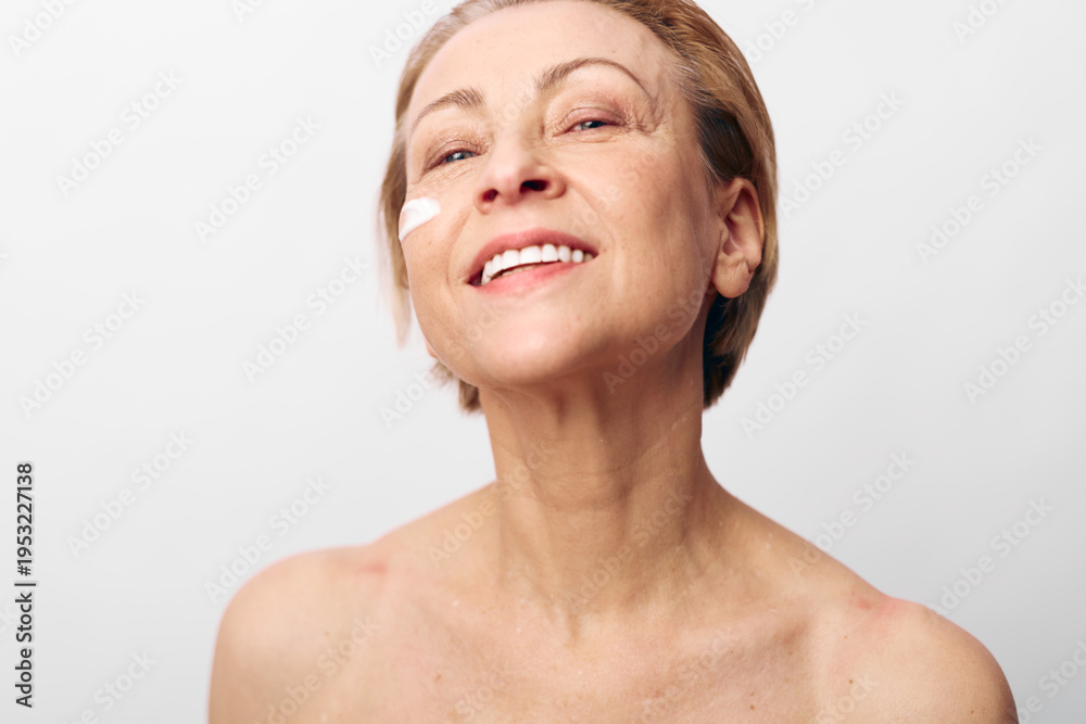 Obraz premium Smiling mature woman with natural skin applies cream on cheek. Close-up on white background. Concept of skincare, aging beauty, self-care, wellness, and healthy lifestyle for women.