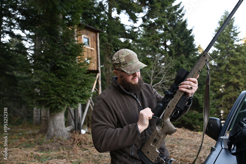 Foto Hunter adjusting scoped rifle near a wooden deer stand in dense evergreen woods