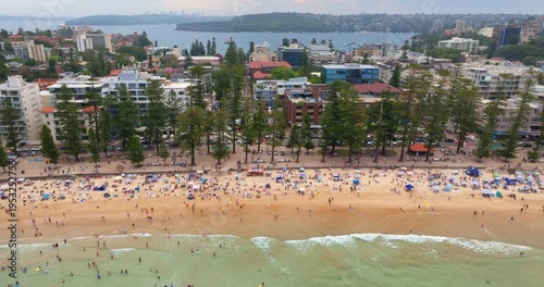Manly Beach in Sydney, Australia, bustling with people enjoying sand and swimming in ocean, showing vibrant coastal lifestyle with surrounding urban development and distant city skyline. Aerial view