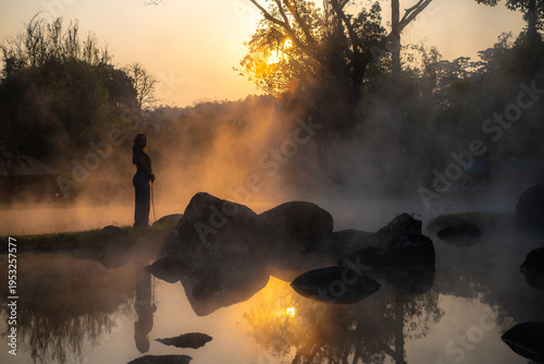 A woman is in a hot spring, with trees behind her, happily bathed in the rising sun.