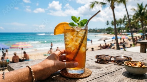 A hand holding a tall glass of iced tea with a straw, garnished with a slice of orange and a sprig of mint, placed on a wooden table with a beach scene in the background.