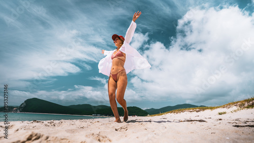 Happy woman enjoying freedom on summer beach, walking barefoot on sand with open arms. Travel lifestyle, joy, relaxation and vacation mood in beautiful coastal landscape. Happy dream vacation