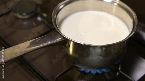 Milk heating in a metal saucepan on a gas stove burner. The process of warming fresh milk in a steel saucepan on an ignited gas burner with a blue flame in a home kitchen