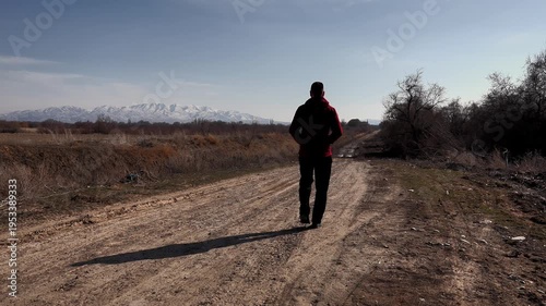 a man walks into the distance along a dirt rural road, against the backdrop of endless rural expanses and snow-capped mountains
