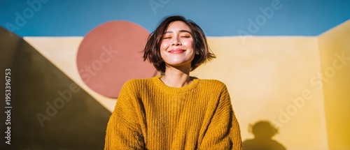 Happy Asian woman smiling with eyes closed wearing a yellow textured sweater against a modern architectural wall for lifestyle advertising and wellness content