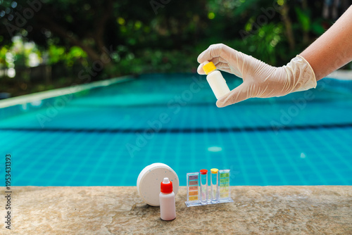 Gloved hand holding reagent bottle above swimming pool with test kit and chlorine tablet on pool edge, water quality control and pool maintenance concept with tropical background