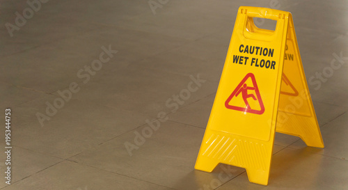 Fotografía A yellow plastic folding sign with a caution wet floor warning placed on a textured grey floor mat to prevent slips and falls in a commercial indoor setting