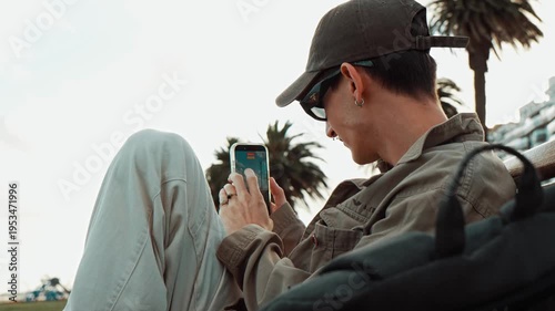 Young man sitting on a park bench between palm trees secretly filming with his mobile phone as if spying or recording something discreetly in an urban outdoor environment