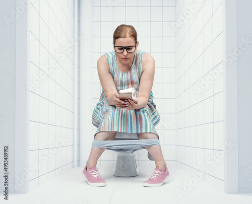 Woman wearing glasses sitting on toilet, holding toilet paper roll and focused on smartphone. Everyday lifestyle moment showing restroom routine and modern mobile phone dependence