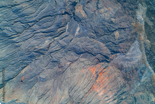 Grey slagheap surface with erosion ridges and orange oxidation, dramatic abstract alien landscape texture, aerial drone photography.