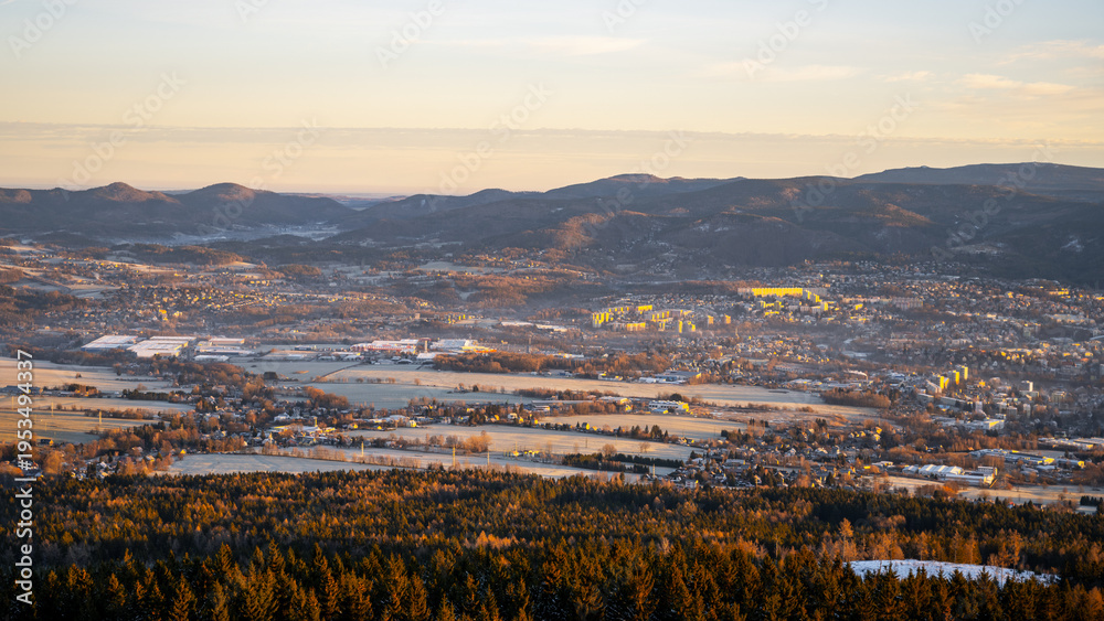 Fototapeta premium A view of Liberec during a winter morning. The landscape shows mountains in the background and fields covered in snow. The town is partly visible with buildings and trees in the foreground.