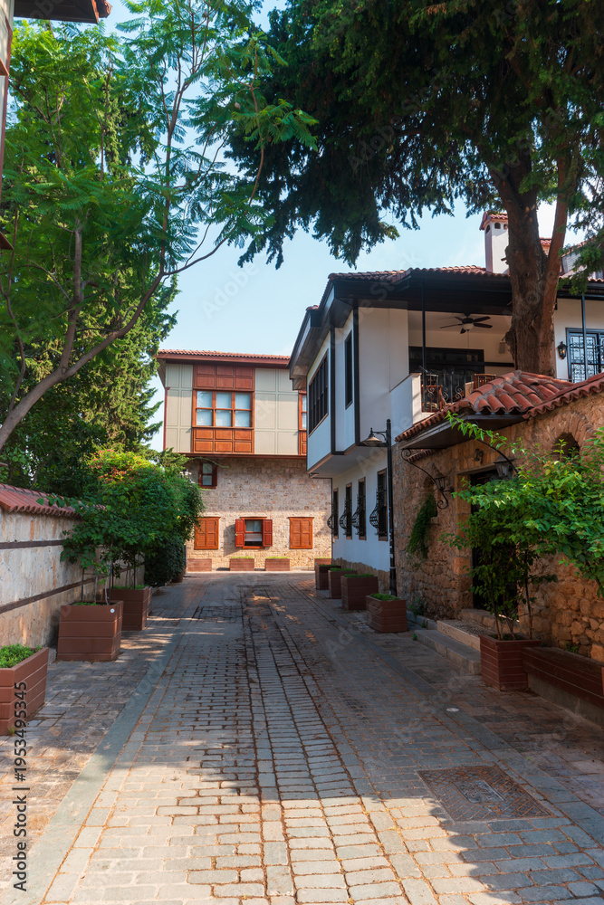 Fototapeta premium Antalya, Turkey. Quiet residential street in Kaleici Old Town with cobblestone pavement, white two-story buildings, red tiled roofs, red door, and trees casting shadows on sunny morning.