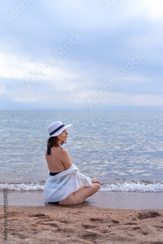Photography A woman in a black and white dress and hat sits on the sand of Lake Issikul, gazing out to sea