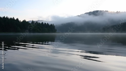 Tranquil lake scene with mist rising from water, surrounded by lush green trees and distant mountains under a soft morning light, creating a serene atmosphere