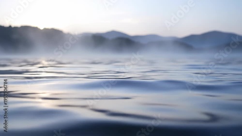 Calm water surface reflecting soft morning light with mist rising from the lake, surrounded by distant mountains under a clear sky at dawn