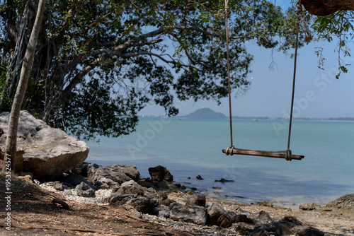 Wooden swing hanging on tree by tropical beach with calm sea and rocky shoreline in Thailand