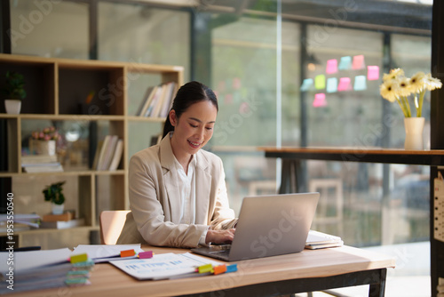 Asian business woman working on her laptop at a desk, smiling with documents and stationary, representing productivity and professional growth in a modern office environment