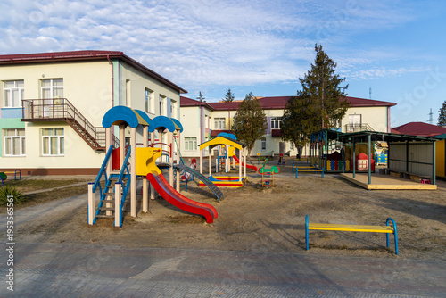 Colorful playground in front of kindergarten building. Wide view of a preschool playground with slides and play structures outside a kindergarten or daycare building on a sunny day.