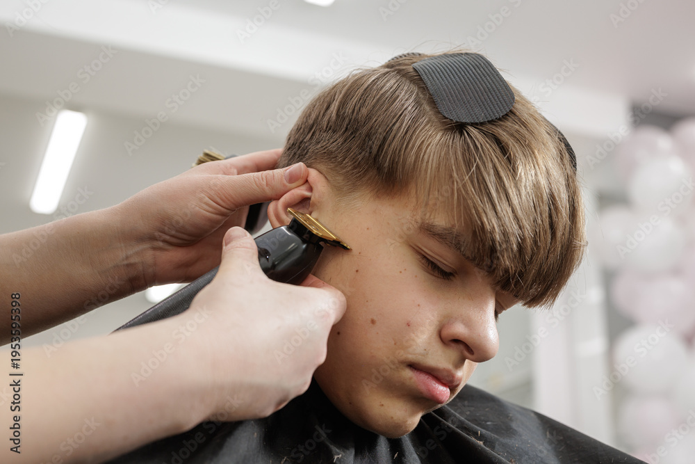Fototapeta premium Hairdresser trimming sideburn with clipper on boy in barbershop