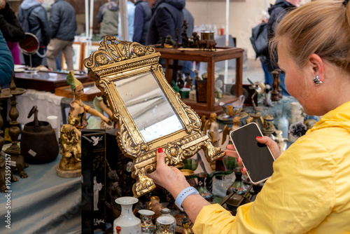 Woman Browsing Antiques at Outdoor Flea Market Holding Ornate Vintage Mirror and Collectibles