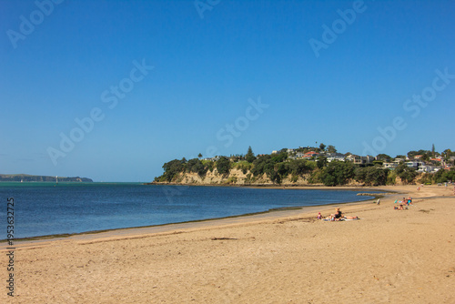 A wide sandy beach stretches along a calm bay with sunbathers relaxing near the shoreline, while houses sit atop a coastal headland beneath a clear blue sky on a warm summer day