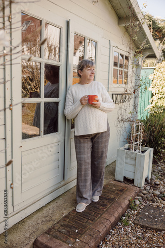 Mature woman standing by house holding mug in garden sunlight