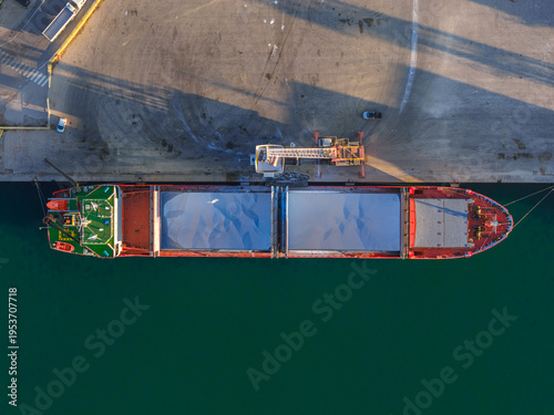 Aerial view of a cargo ship docked at the port, its red hull contrasting with the teal water and the pale concrete of the pier, Vasto, Abruzzo, Italy.