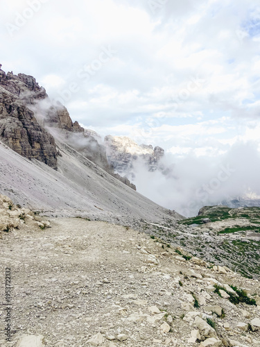 A wide shot of a rugged mountain landscape featuring a steep rocky scree slope, jagged peaks shrouded in mist, and a gravel hiking trail in the foreground.