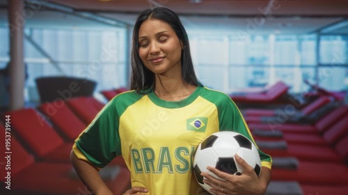 Woman in yellow brasil jersey holding soccer ball with hand on hip, smiling toward camera in airport lounge; team pride.