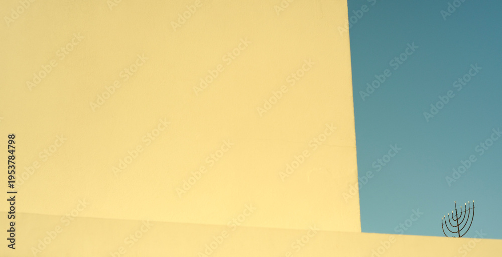Obraz premium Minimalist composition of a geometric yellow wall edge against a blue clear sky, with a menorah silhouette on the roof.