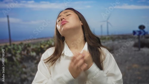 Young hispanic woman in white shirt clenches fists before windmill towers in field under blue sky; strength resilience perseverance determination.