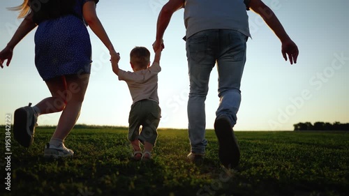 Parents lift child while running on grass field at sunset outdoors. Family swings boy between them. Mother and father lift child jumping. Parents run with boy on field. Dog watches family playing.