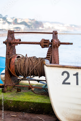 Konstfotografi Rusting fishing winch on a British shingle beach with small boat, capturing a quiet coastal scene in a traditional fishing town
