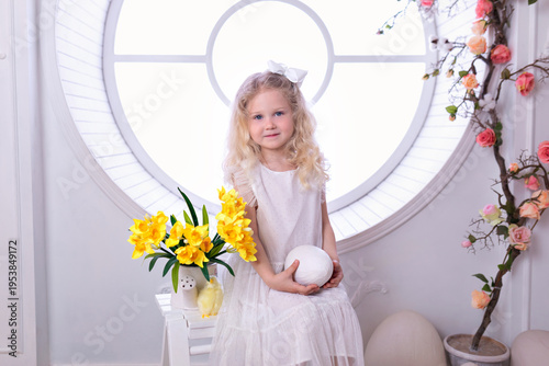 Wallpaper Mural A little curly blonde girl in white dress with with large Easter eggs sit on chair with spring interior decoration room. Happy smile child girl holding an easter egg in her hands near big window Torontodigital.ca