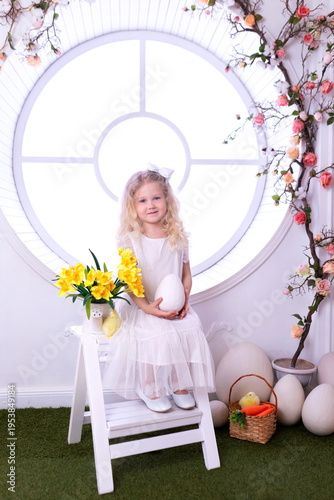 Wallpaper Mural A little curly blonde girl in white dress with with large Easter eggs sit on chair with spring interior decoration room. Happy smile child girl holding an easter egg in her hands near big window Torontodigital.ca