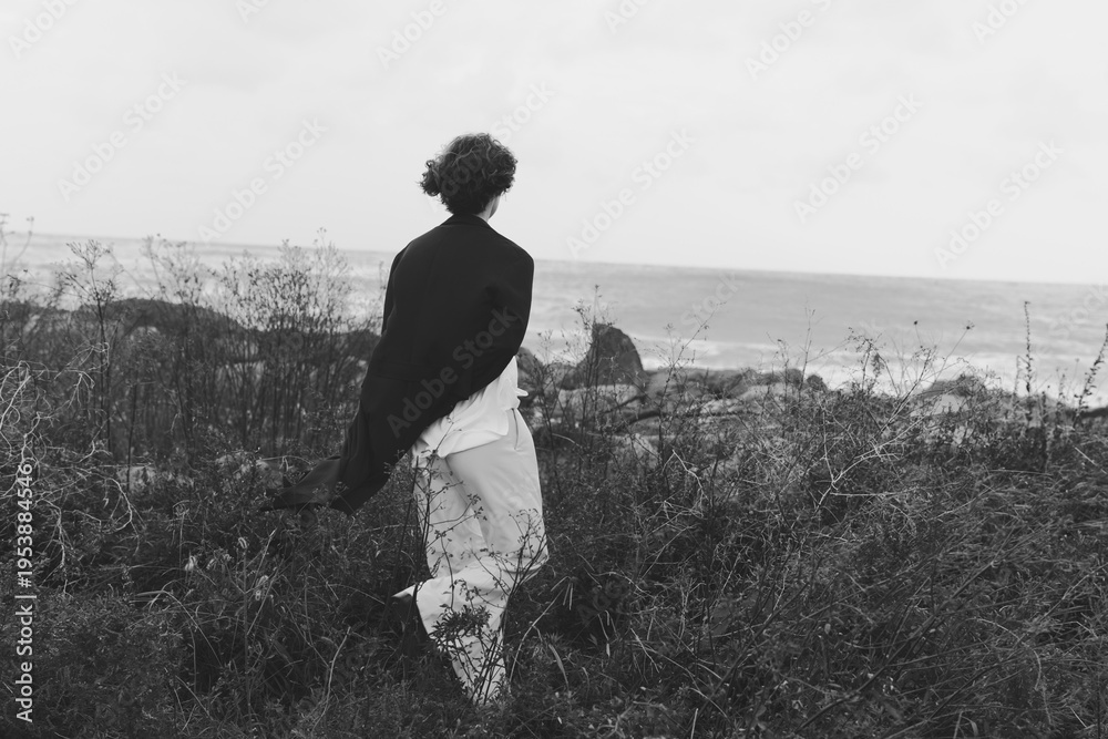 Fototapeta premium Silhouette of a woman walking through meadow near coast in grayscale, wind in hair, tranquil and solitary journey amid sea spray and wildflowers