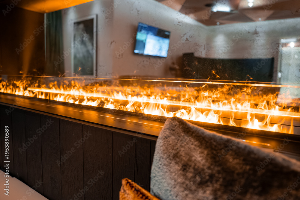 Fototapeta premium A linear fireplace flickers behind glass in a modern alpine lounge in Zermatt, Switzerland. Warm amber light plays on cushions, dark wood, and a wall mounted TV.