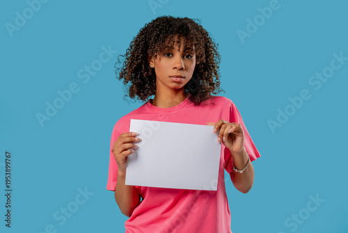 Serious woman holding blank a4 white sheet of paper on blue studio background.Protest, complaint, announcement, message, frustration, advertising mockup, copy space for text or design