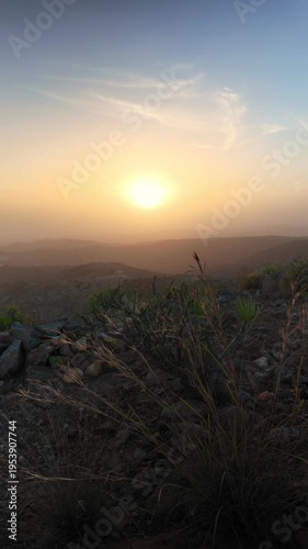 Golden sun setting over a hazy mountain landscape in the Canary Islands, with arid vegetation and rocks in the foreground creating a serene and dusty atmosphere during a beautiful summer evening