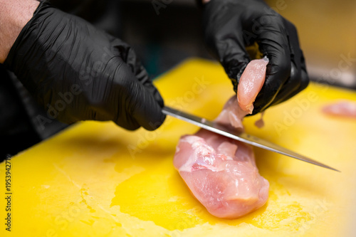 фотографія Chef in black gloves slicing raw chicken breast with a sharp knife on a yellow cutting board in a professional kitchen