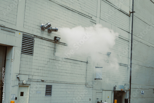 Foto Smoke billows from vents on a factory wall during daytime in an industrial area