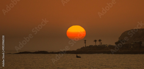 The setting sun descends over the Atlantic Ocean towards the cliffs as people play in the sea and on the beach at the end of another hot day in Taghazout, Agadir, Morocco