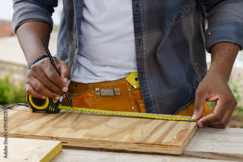 African American man measuring plywood with yellow tape measure on bench, wearing tool belt