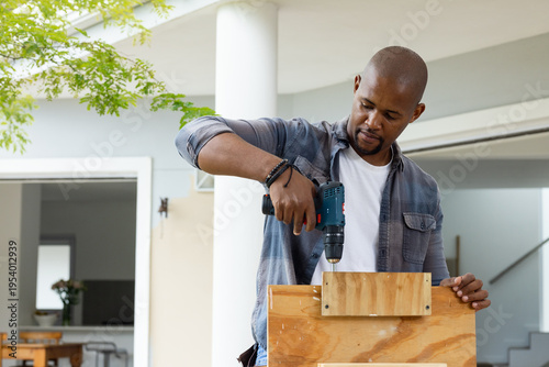 African man using cordless drill while fastening wooden faceplate to plywood on front porch