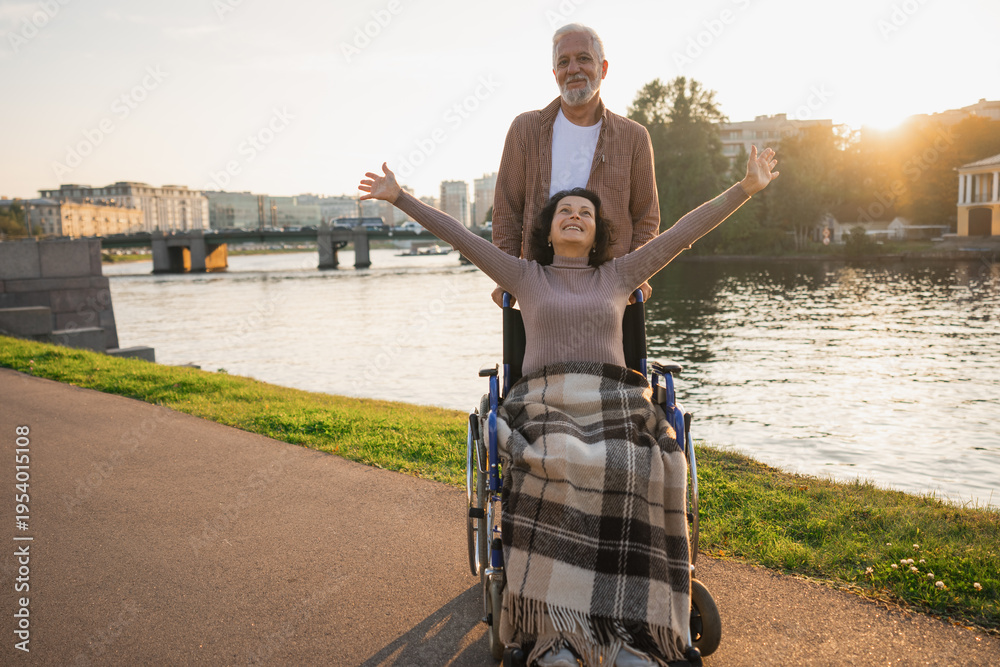 Fototapeta premium Senior woman in wheelchair walking with caregiver old man on road. Family couple man husband supporting taking care of paralyzed woman wife in chair for people with disability outdoor. Rehabilitation