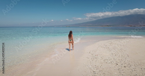 Sexy long hair woman in bikini walking barefoot on idyllic white sand bar leading into crystal clear turquoise water, vacation on tropical paradise, Gili Islands with a distant volcano. Back view