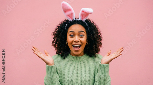 Joyful young woman with curly hair wearing charming pink bunny ears and a green sweater, displaying an excited, surprised expression with open hands and a wide smile.